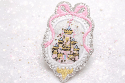 Princess castle shaker embroidery patch featuring a glitter silver scalloped frame, pink bow detail, and embroidered fairy tale castle with loose sparkle confetti inside. This handmade princess castle appliqué patch is shown isolated on a white background and is designed for kids clothing, dresses, backpacks, jackets, and craft projects.