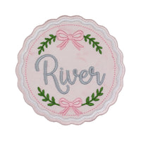 Blush pink embroidered wax seal shaped patch with a wavy pink and white satin stitched border, pink bows at the top and bottom, green leafy sprigs, and the name River in gray script across the center.