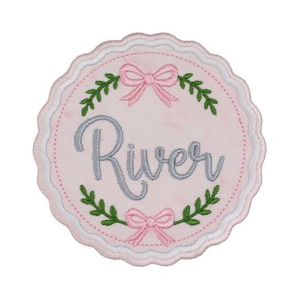 Blush pink embroidered wax seal shaped patch with a wavy pink and white satin stitched border, pink bows at the top and bottom, green leafy sprigs, and the name River in gray script across the center.