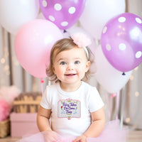 Child in a white shirt with a fairy-themed design, surrounded by pink and purple balloons.
