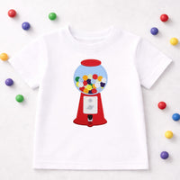 Gumball machine embroidery patch sewn onto a white children’s t-shirt, featuring a red vintage gumball dispenser filled with colorful embroidered gumballs inside a blue globe. This retro candy machine sew on patch is shown on kids clothing to demonstrate use on shirts, jackets, backpacks, and handmade apparel.